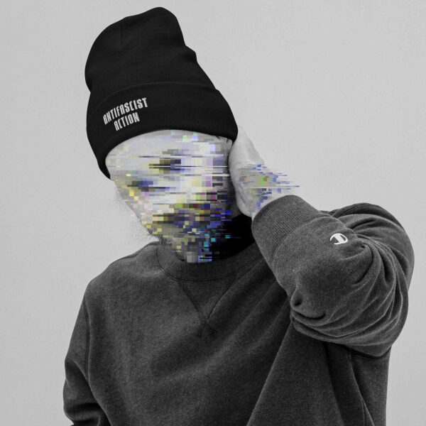 A man putting his hand to his neck, wearing a pullover and a Beanie with an embroidery in the lower front, saying 'Antifascist Action', his face is made unrecognisable with colourful glitches