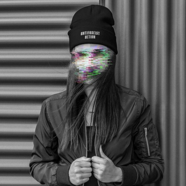 A woman standing in front of a corrugated iron wall, wearing a Beanie with an embroidery in the lower front, saying 'Antifascist Action', her face is made unrecognisable with colourful glitches