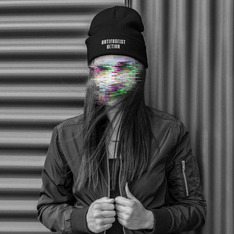 A woman standing in front of a corrugated iron wall, wearing a Beanie with an embroidery in the lower front, saying 'Antifascist Action', her face is made unrecognisable with colourful glitches