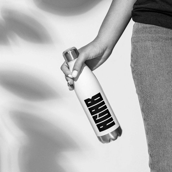 A female right hand, holding a white bottle with a black 'ACAB' or 'All Cops Are Bastards' print on it, with brushed steel lid and bottom, the womans right leg is partzly in the frame, shadow of a large flower on the white wall in the background