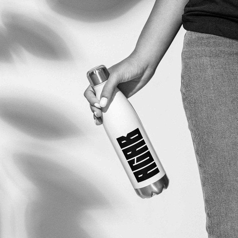 A female right hand, holding a white bottle with a black 'ACAB' or 'All Cops Are Bastards' print on it, with brushed steel lid and bottom, the womans right leg is partzly in the frame, shadow of a large flower on the white wall in the background