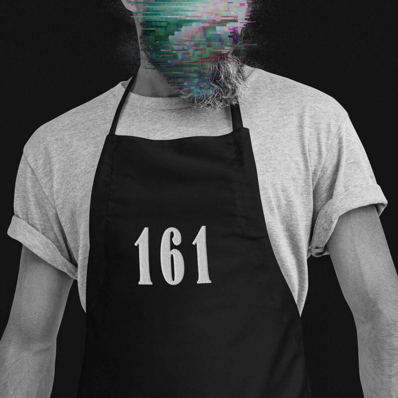 Fashionable, beardy man wearing an apron against racism, which has an embroidery in the middle saying '161' in large serif digits, which is short for 'antifascist action'.