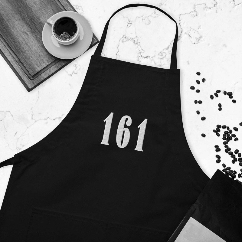 Anti-racist apron with the embroidery '161', which is short for 'antifascist action', next to a coffee mug on a wooden chopping board and some coffee beans on a marvel floor or table