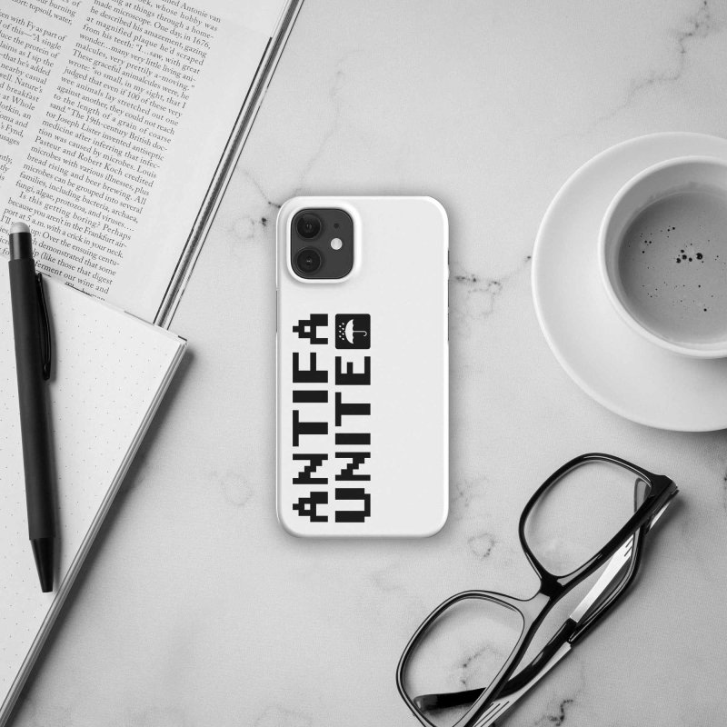 Back of an iPhone with a white phone case, located on a white marvel surface, the case is saying 'antifa unite' in black letters, with a little icon showing an umbrella under water droplets, white on black square with rounded corners, to the right of the phone there is a brochure, a dotted paper notebook and a black ballpoint pen, to it's left there is a white coffee mug with coffee and black glasses to it's lower right